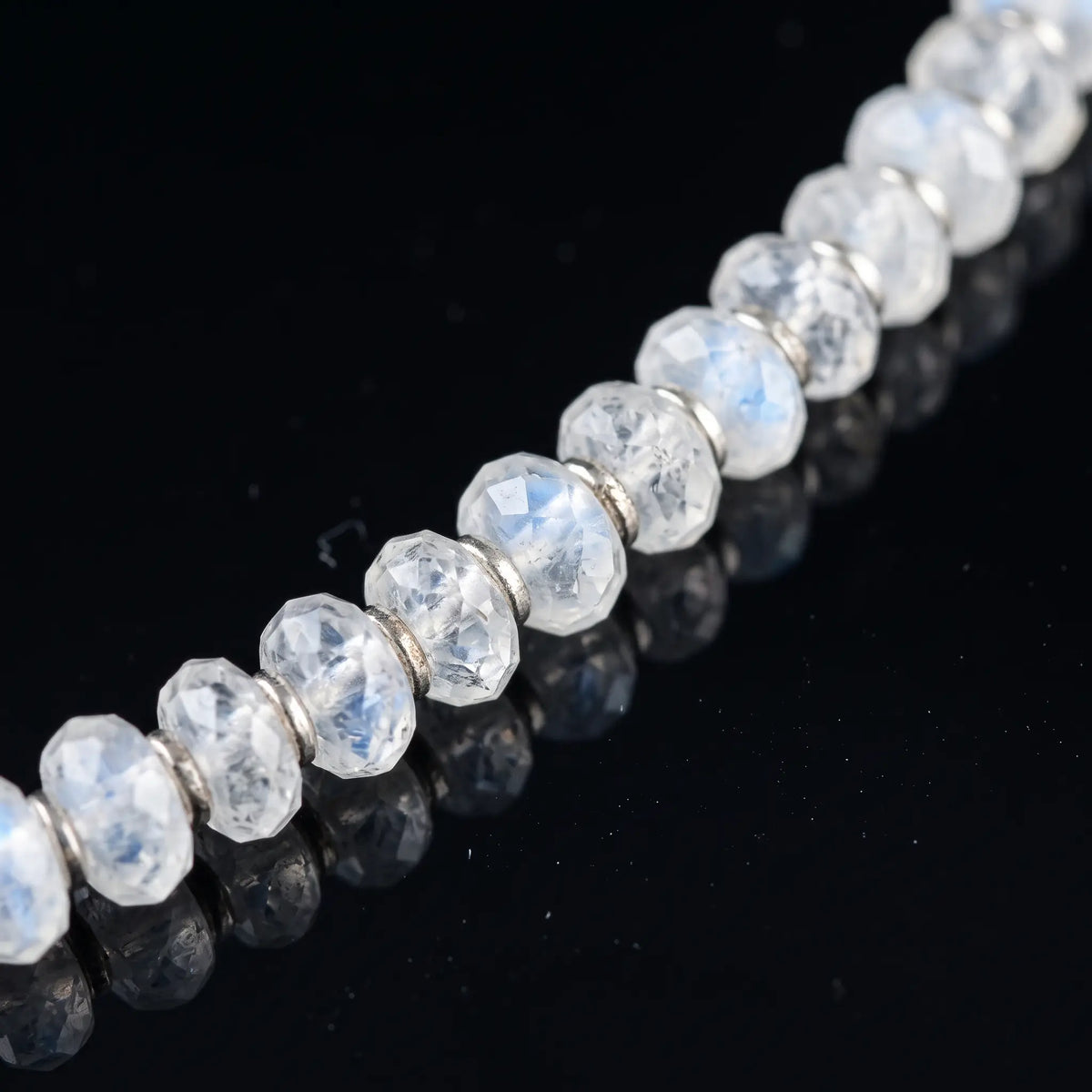 Close-up of Moonstone Bracelet X (4mm), showing pale faceted moonstone beads with blue flashes on a black reflective surface.