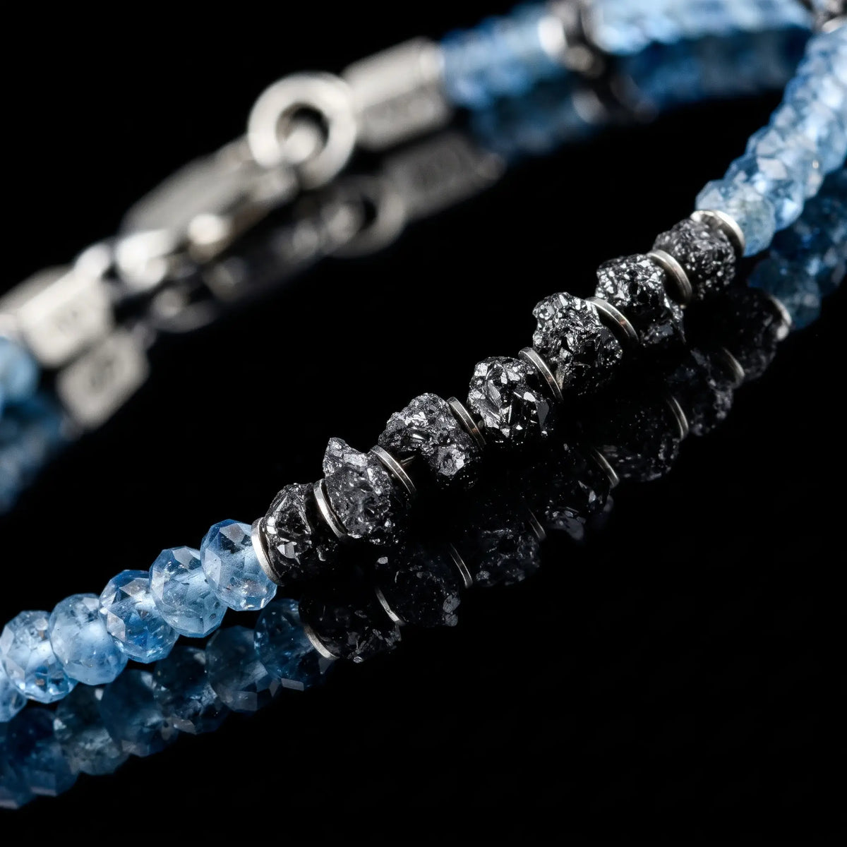 Close-up of Raw Black Diamond - Aquamarine Bracelet I (4mm), showing its rough black diamond centre and faceted blue aquamarine on a black reflective surface.